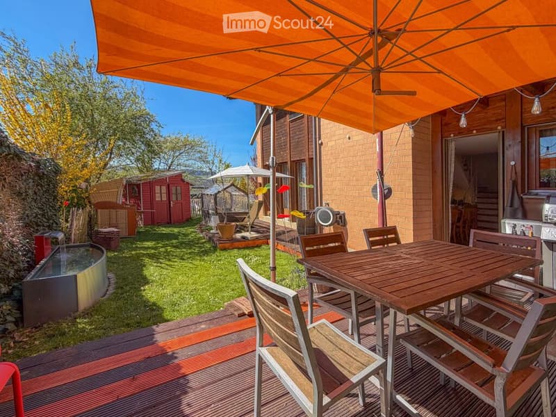 Outdoor patio with wooden table and chairs, large umbrella, small fountain, and surrounding greenery.
