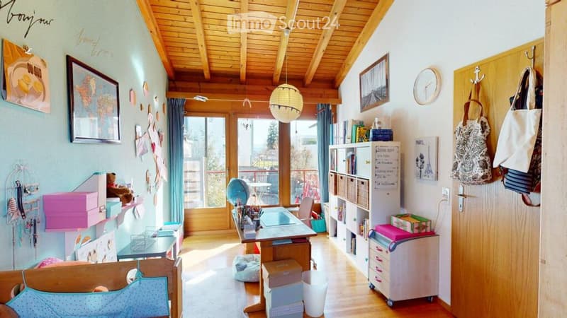 A cozy, well-lit room with wooden ceilings and floors, featuring a desk with a globe, a bookshelf, a storage unit, and various decorative items including picture frames and a clock. The room has large windows with curtains and a door leading to a balcony.