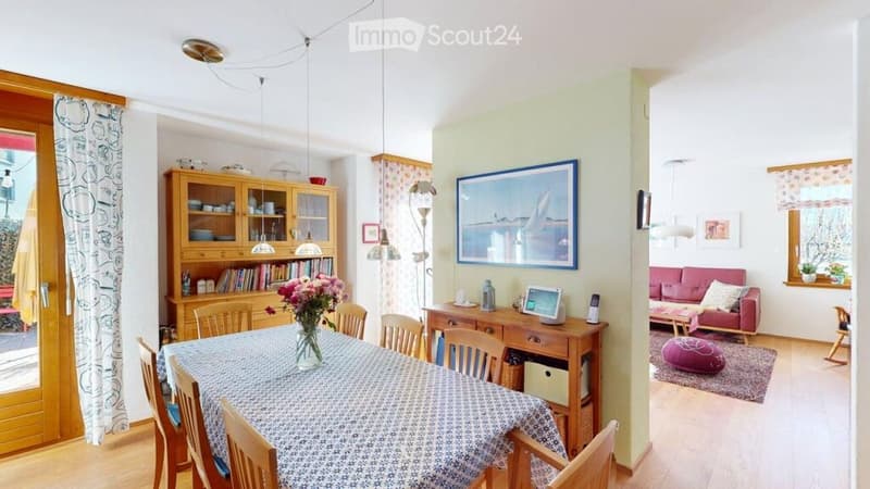 Bright living and dining area with wooden flooring, a red sofa, a dining table with chairs, a wooden cabinet, and a large picture frame on the wall.