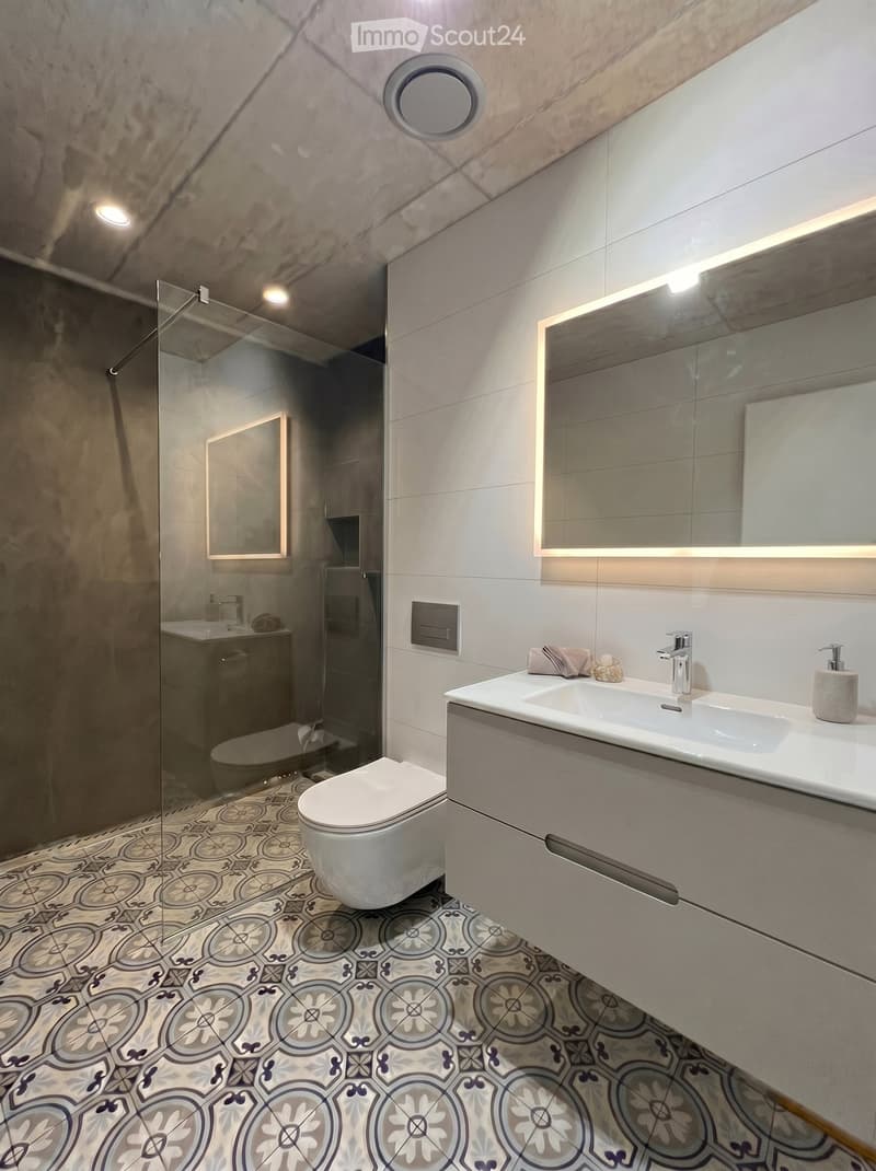 Modern bathroom with a white sink, chrome faucet, large mirror with built-in lighting, toilet, glass-enclosed shower, and decorative patterned tiles.