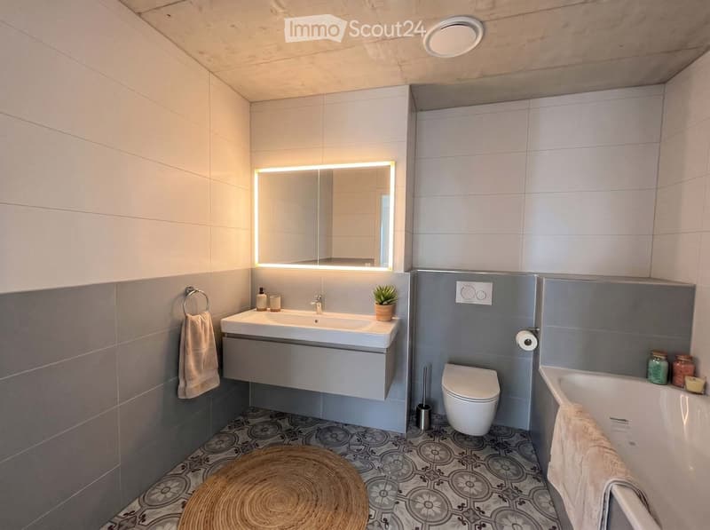 Modern bathroom with a white sink, mirror with LED lighting, toilet, bathtub, and decorative elements including a round woven stool and small plant.