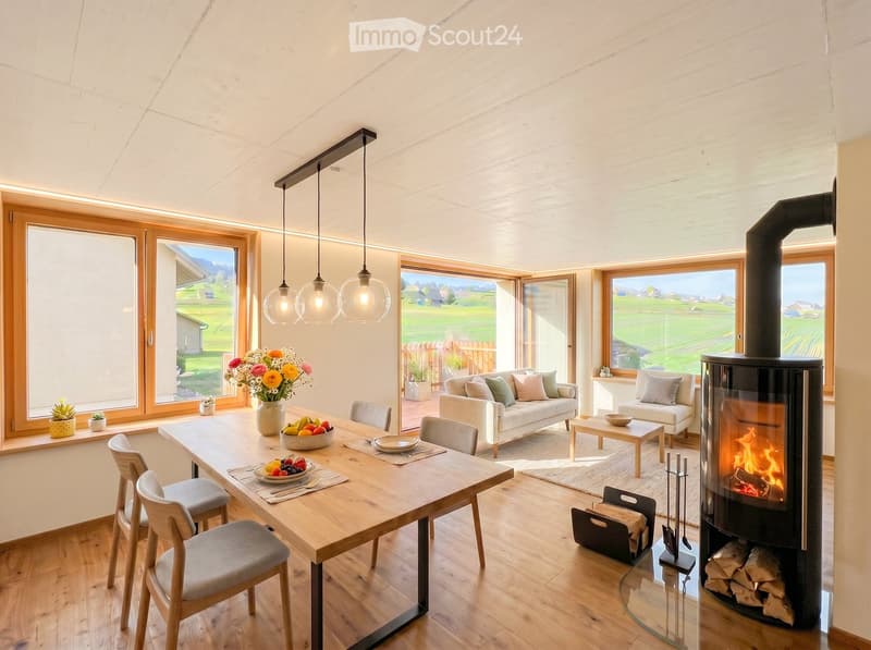 Bright living and dining area with large windows overlooking a green landscape, featuring a sofa, coffee table, dining table with chairs, and a modern wood-burning stove.