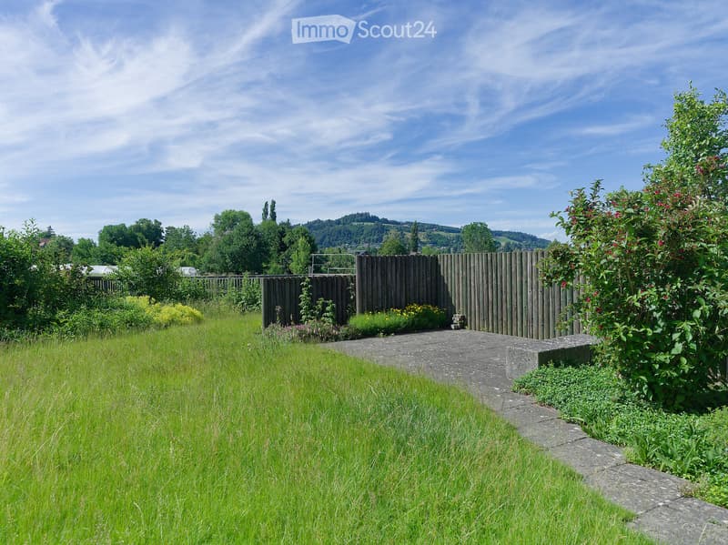 A well-maintained garden with a wooden fence, lush green grass, and various flowering plants, set against a backdrop of trees and hills under a clear blue sky.