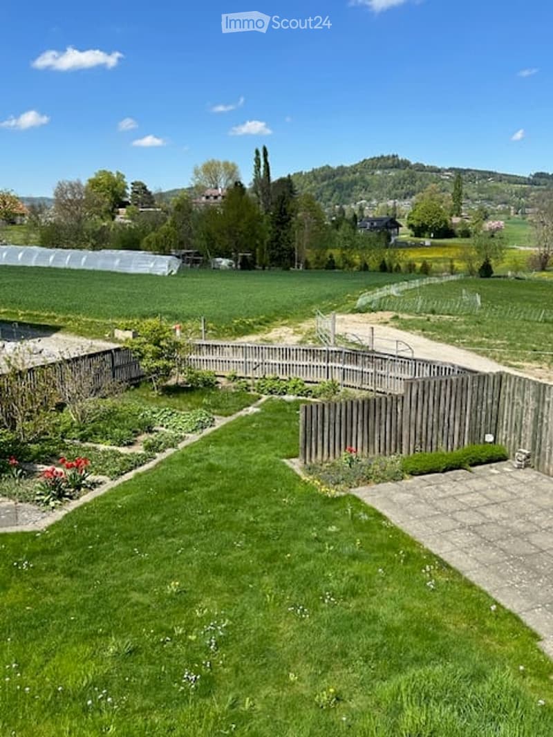 Well-maintained garden with neatly trimmed grass, flower beds, wooden fence, and a greenhouse in the background. The area is surrounded by a wooden fence and features a gravel pathway.