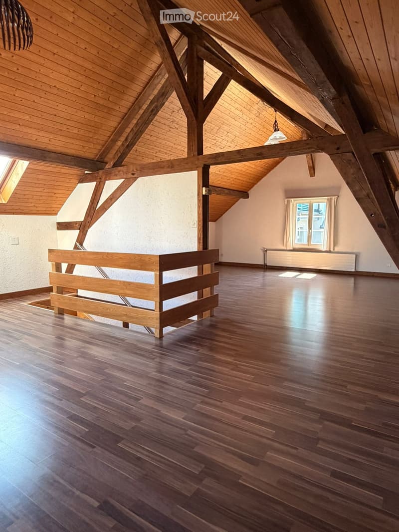 Empty attic space with wooden beams, wooden floor, and large windows with curtains. No furniture or equipment present.