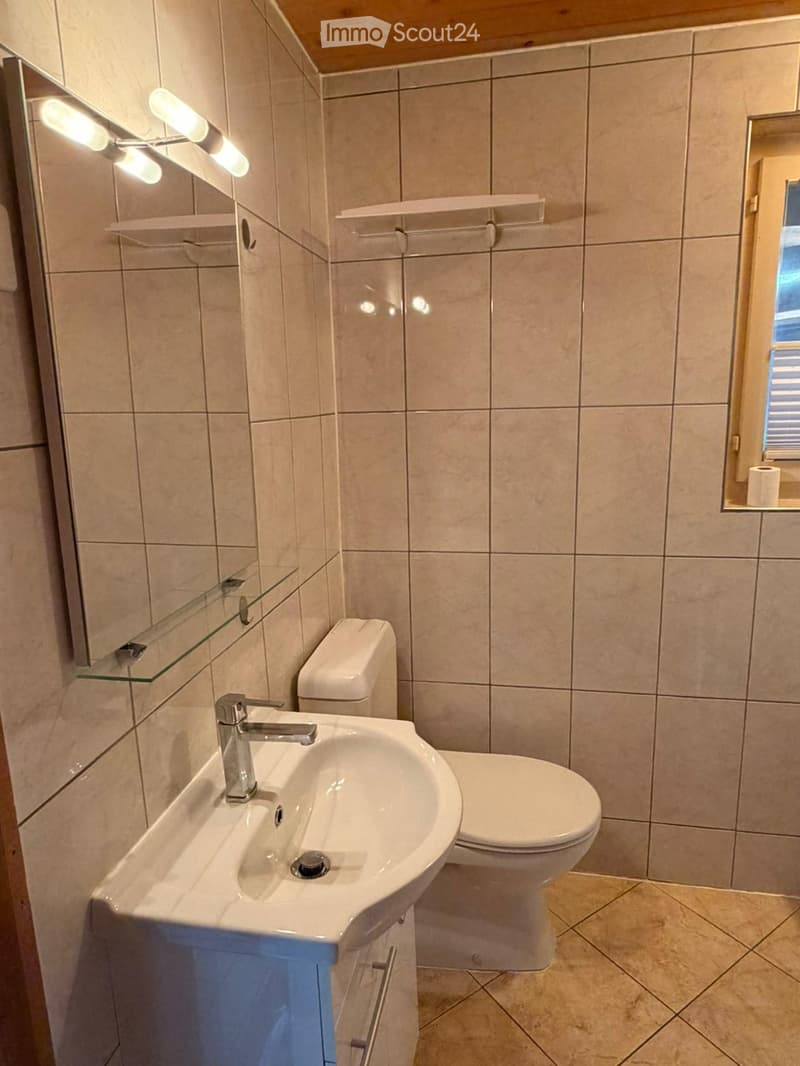 Modern bathroom with beige tiled walls and floor, white sink with chrome faucet, toilet, mirrored cabinet with glass shelves, and a window with blinds.
