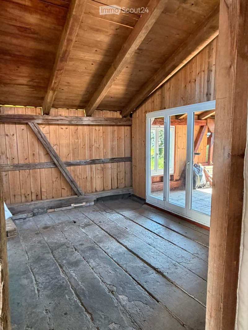 An empty attic space with wooden beams, exposed wooden walls, and a stone floor. A large sliding glass door leads to an outdoor area.