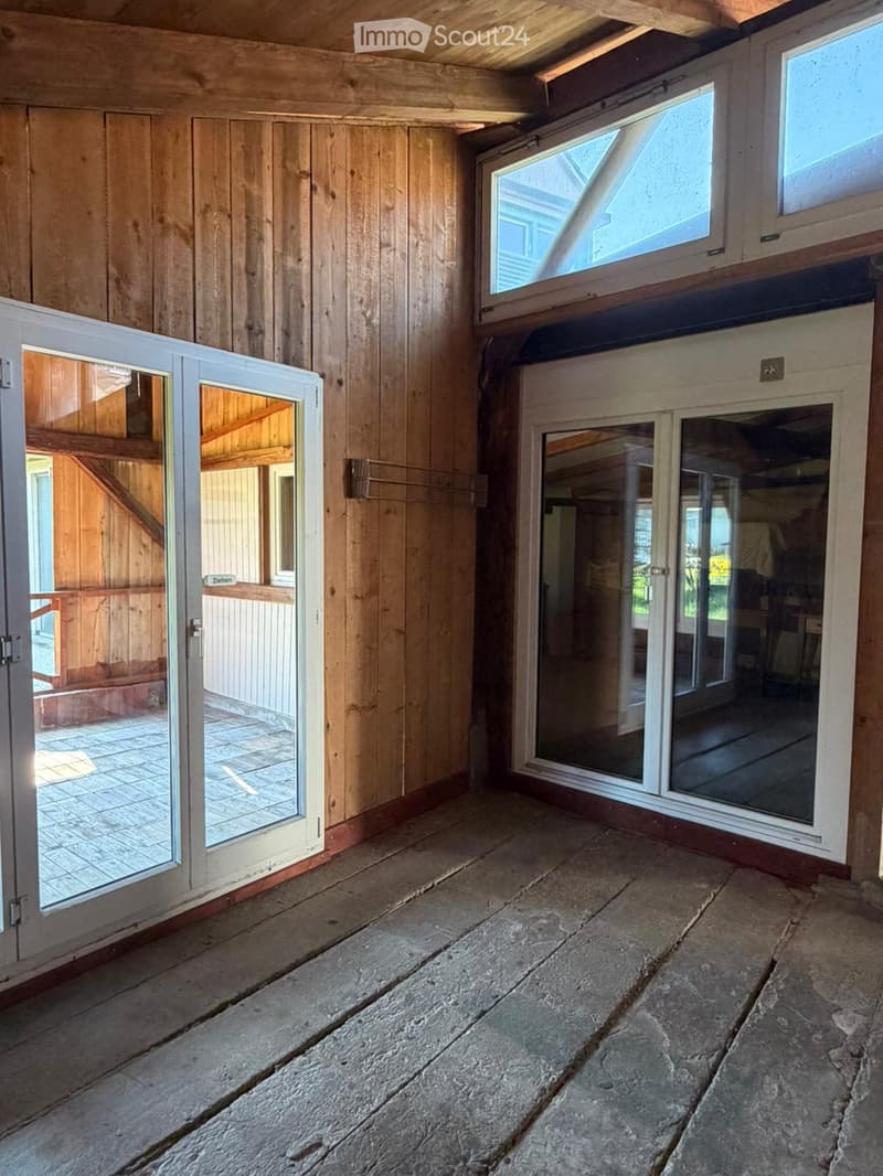 Empty room with wooden walls, stone floor, and large windows with white frames. The space has a rustic, unfinished look with visible wooden beams on the ceiling.