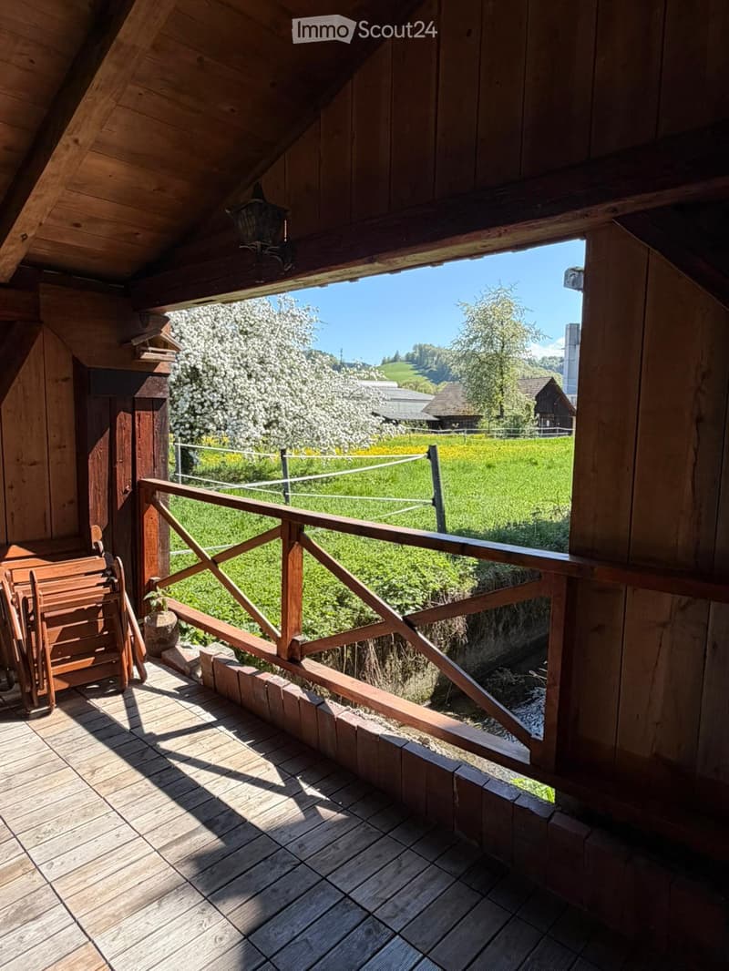 Wooden balcony with railing, wooden floor, and a view of a green field and trees.