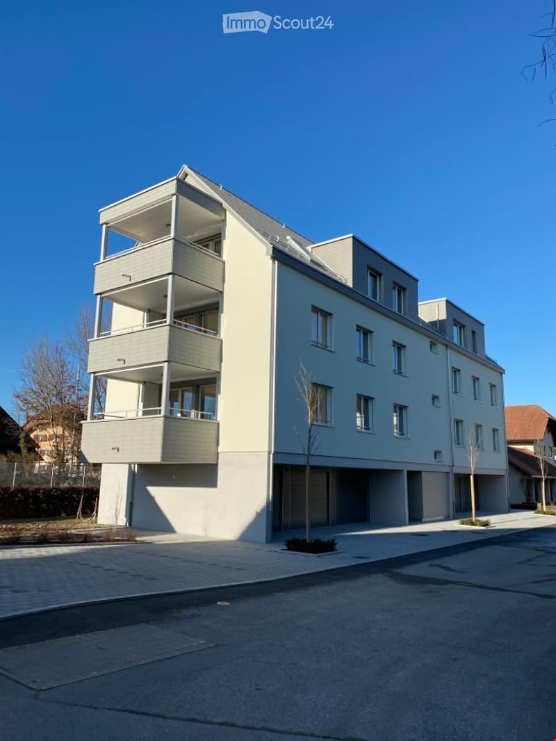 Modern multi-story residential building with balconies and large windows, surrounded by a paved area and sparse landscaping.