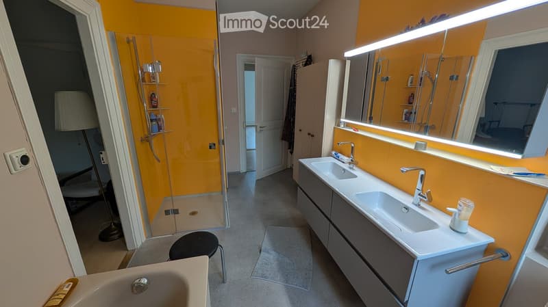Modern bathroom with double sink, large mirror, bathtub, and a glass-enclosed shower area. Features include a grey cabinet, a black stool, and a floor lamp.