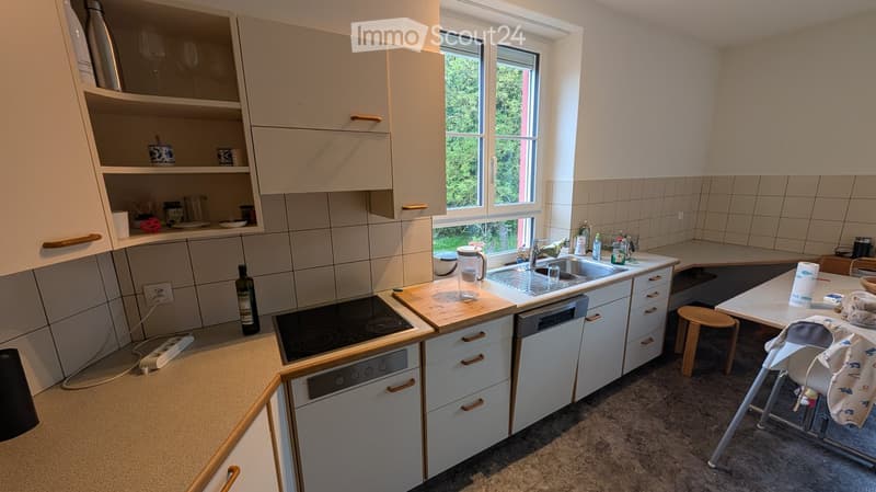 Modern kitchen with white cabinets, stainless steel appliances, and a large window overlooking greenery. Includes a sink, stove, dishwasher, and a small table with a chair.