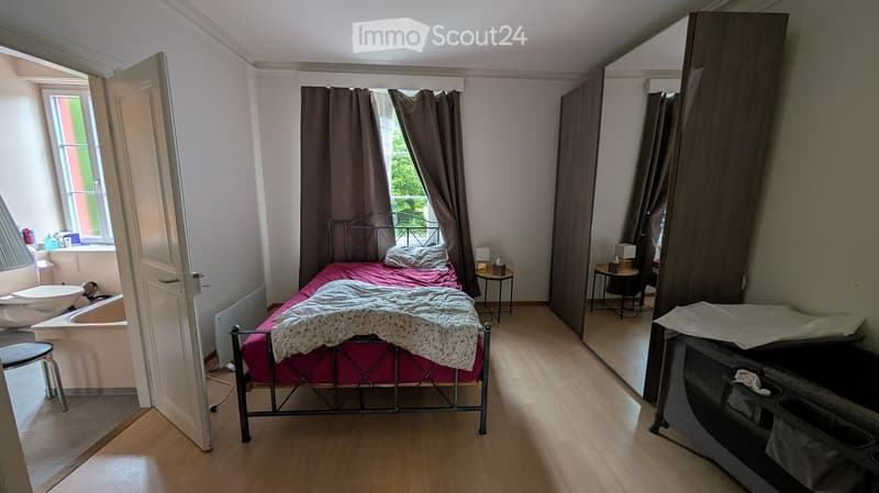 A furnished bedroom with a metal-framed bed, pink bedding, a nightstand, a mirror, a wardrobe, and a small bathroom area visible through an open door.