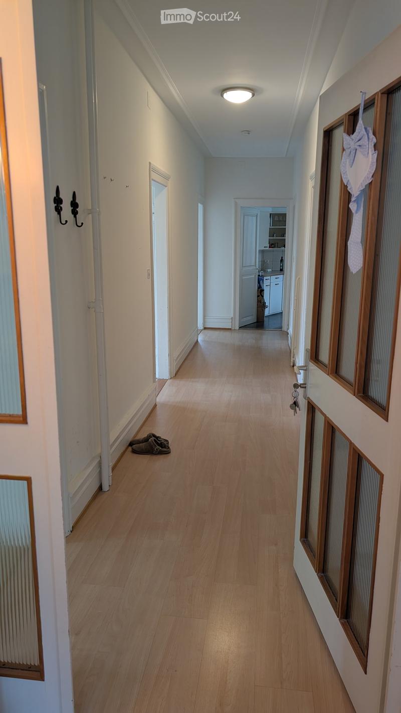 A long, narrow hallway with light wooden flooring, white walls, and several doors. A pair of slippers is placed near the left wall, and a small decorative item hangs on the right wall.