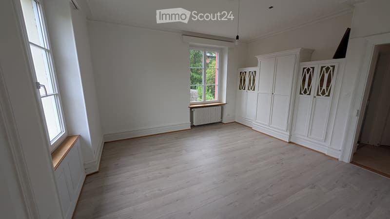 Empty room with light wooden flooring, white walls, built-in white cabinets, and large windows allowing natural light.