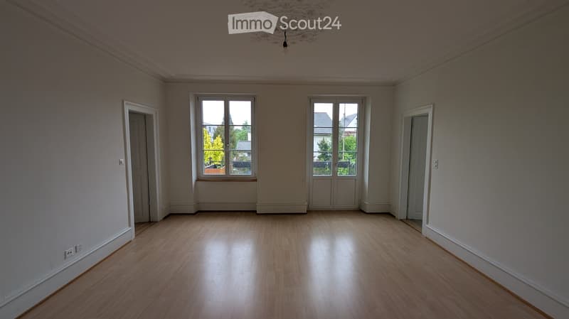 Empty room with light wooden flooring, white walls, and large windows with white frames. The room has two doors and a chandelier hanging from the ceiling.