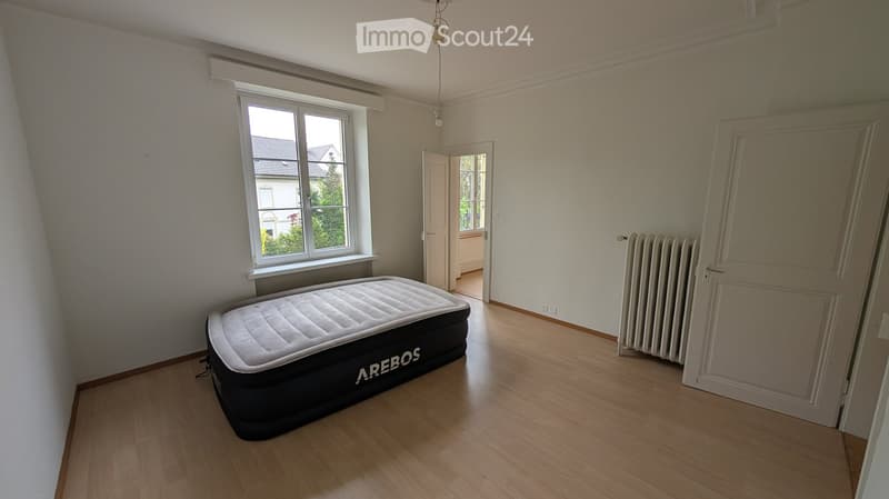 Empty room with wooden flooring, a single air mattress, a radiator, and a window with a view of a neighboring house.