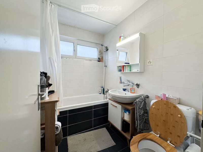 Bathroom with a bathtub, sink, toilet, mirror, and wooden toilet seat. Includes a cabinet, towel, and various toiletries.
