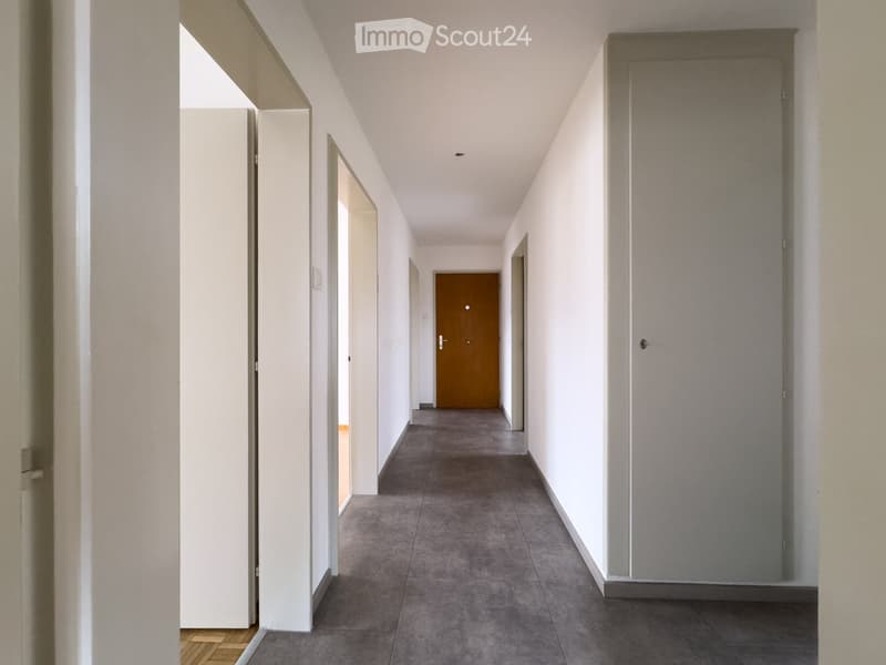 Empty hallway with white walls, grey tiled floor, and several doors including a wooden door at the end.