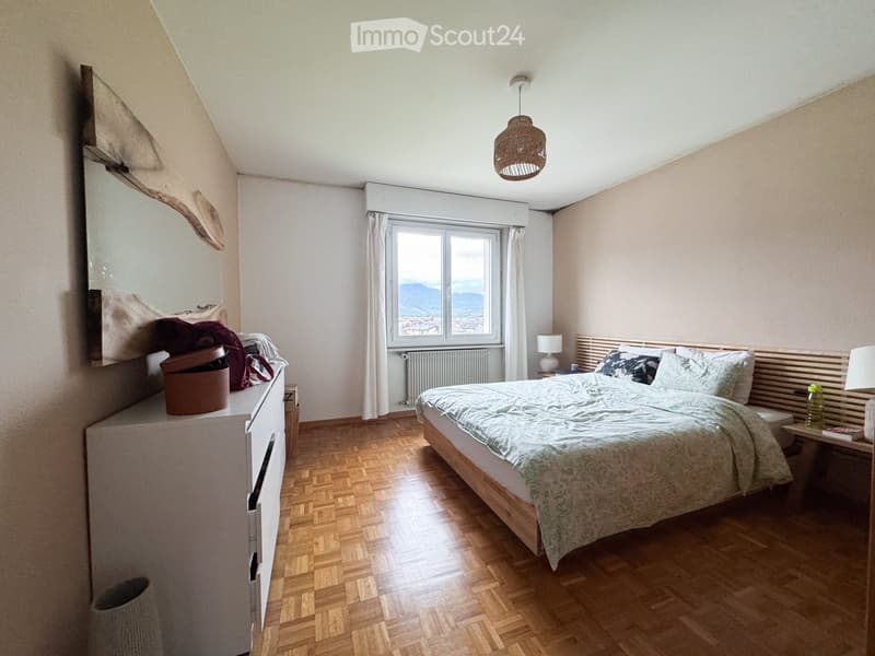 A cozy bedroom with wooden flooring, a large window offering a scenic view, a neatly made bed with pillows and a duvet, a nightstand with a lamp, a mirror, and a drawer with some items on top.