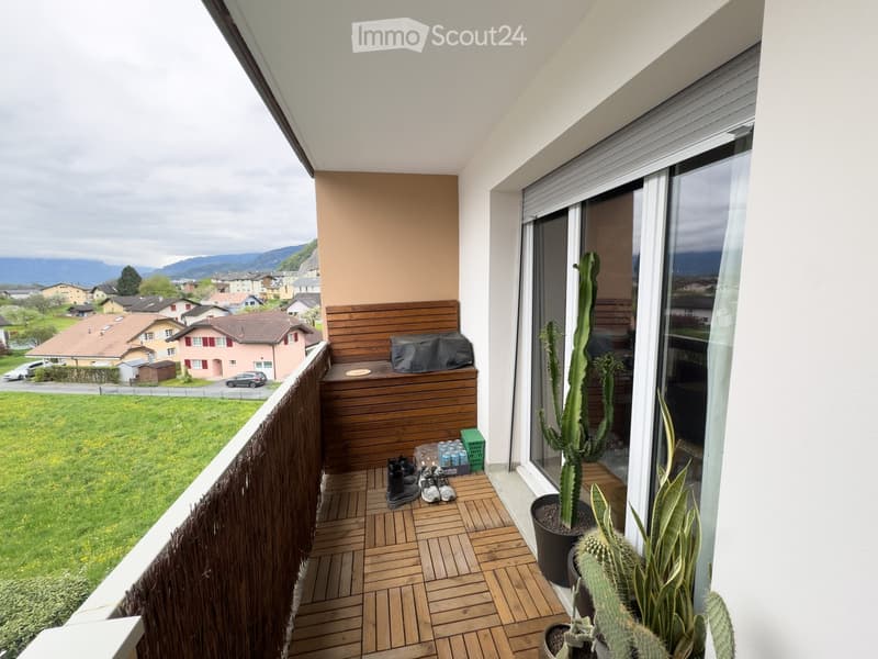 Balcony with wooden decking, potted cacti, a wooden bench, and shoes placed near the railing. Offers a scenic view of a green landscape and distant mountains.