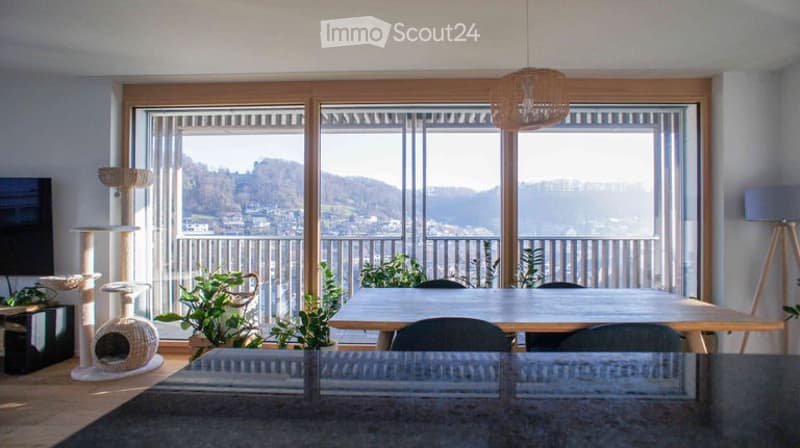 Bright living room with large windows offering a scenic view, featuring a cat tree, a dining table with chairs, and a hanging lamp.