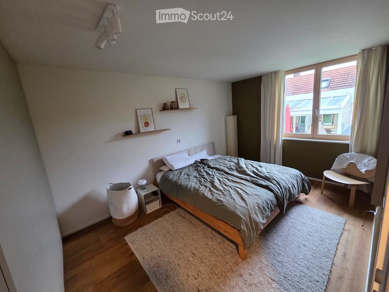 Cozy bedroom with wooden flooring, a bed with grey bedding, a nightstand with a lamp, a small chair, and a shelf with decorative items.