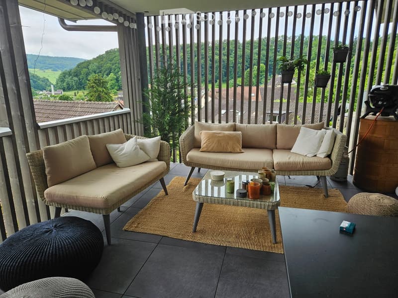 Balcony with a beige wicker sofa, glass coffee table, and two round stools, surrounded by potted plants and a scenic view.
