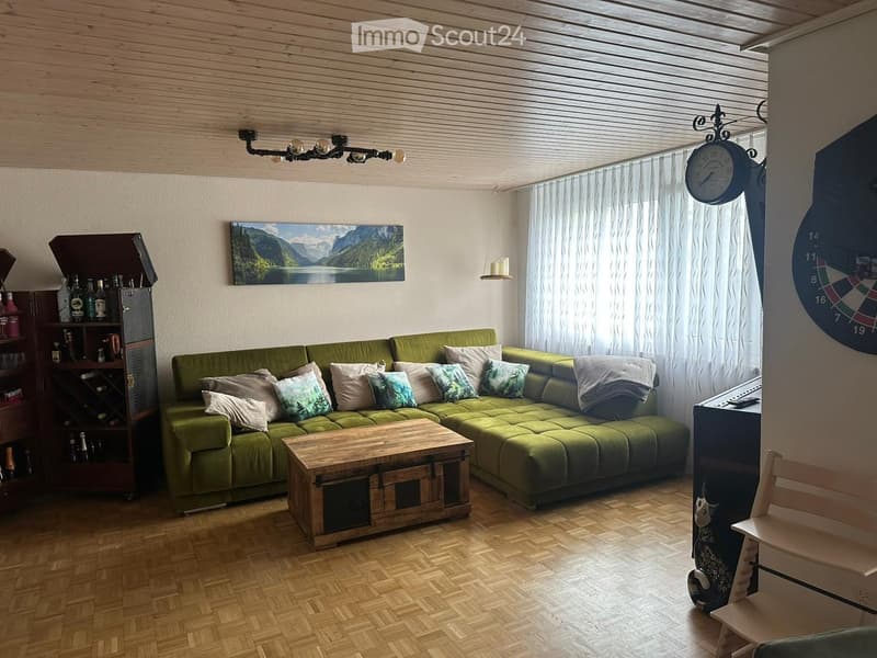Living room with green sectional sofa, wooden coffee table, wine rack, painting, clock, and a chair near the window.