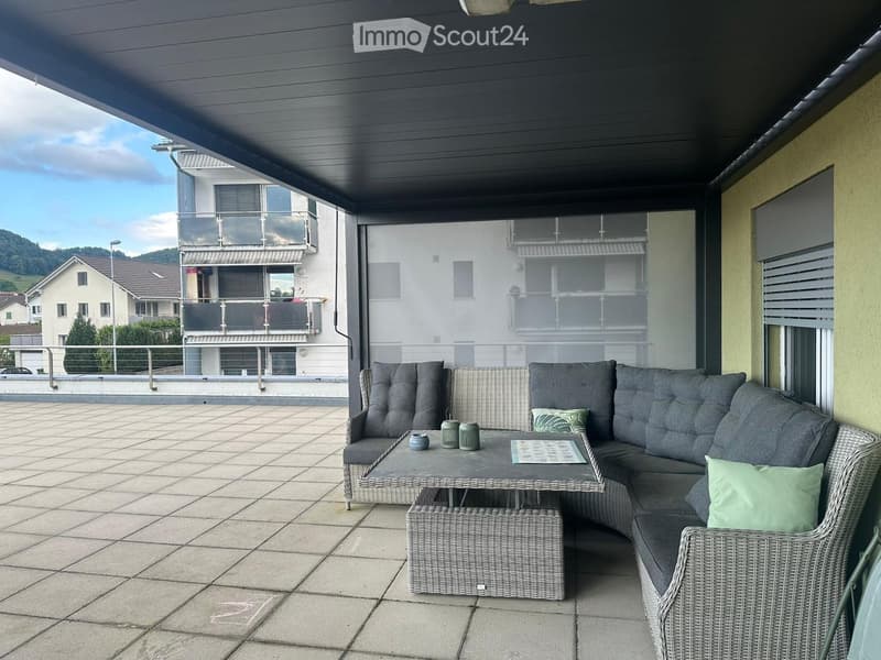 Balcony with a grey wicker sofa, a small table, and two stools, featuring a scenic view of the surrounding area.