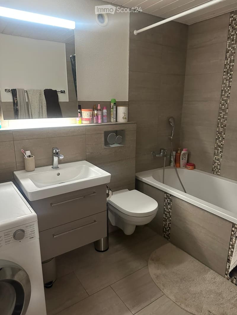 Modern bathroom with a sink, mirror, towel rack, toilet, bathtub, and showerhead. The sink has a modern faucet and a small cabinet underneath. The bathtub has a tiled border and a showerhead attached to the wall. A towel rack with towels is visible above