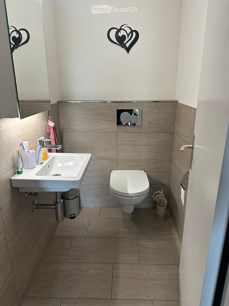 Modern bathroom with a white sink, chrome faucet, toilet, and a mirror above the sink. Beige tiles on walls and floor, with a decorative heart-shaped wall art.