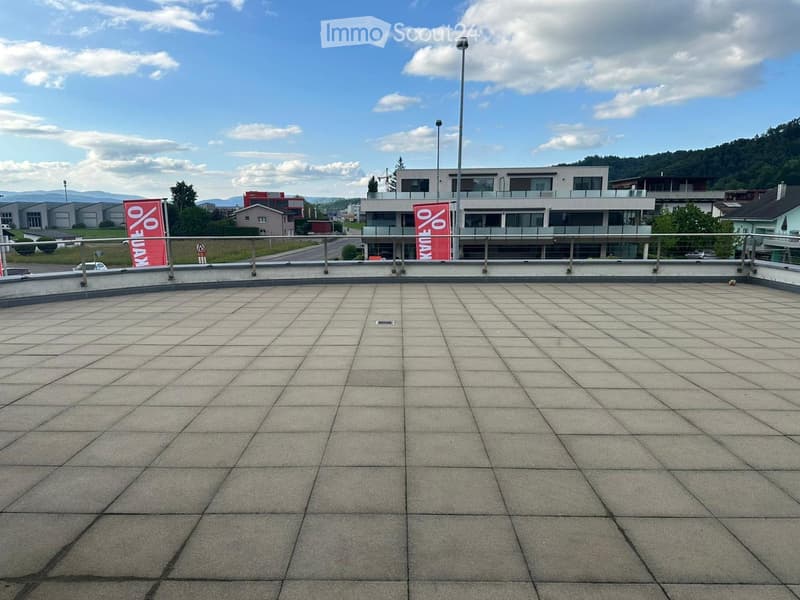 Empty rooftop terrace with tiled surface, metal railing, and distant buildings and mountains in the background.