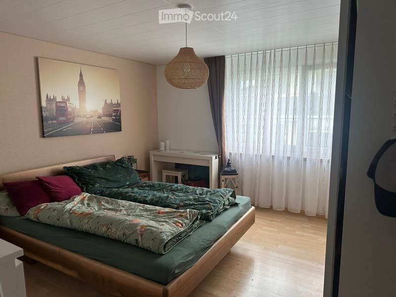 Cozy bedroom with a wooden bed, green duvet, red pillows, a small desk, and a hanging woven lamp.