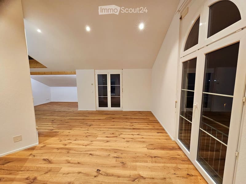 Empty room with wooden flooring, white walls, and large windows with glass doors leading to another space.