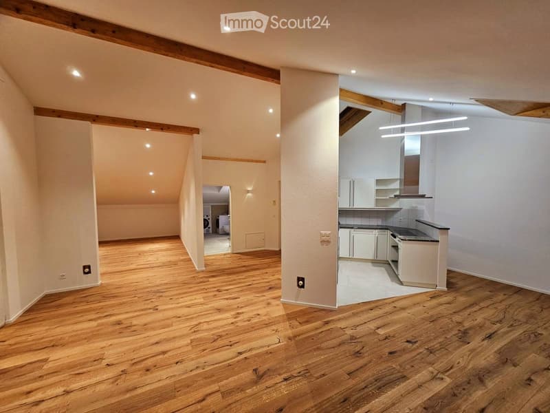 An unfurnished room with wooden flooring, white walls, and exposed wooden beams on the ceiling. The room features a small kitchen area with white cabinets and a countertop, and a separate laundry area with a washing machine and dryer visible in the backgr