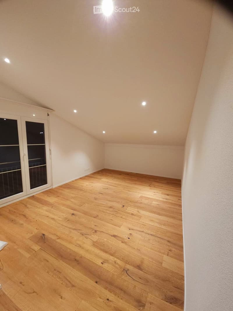 Empty room with wooden flooring, white walls, and recessed ceiling lights. Features a large window with a view of a balcony.