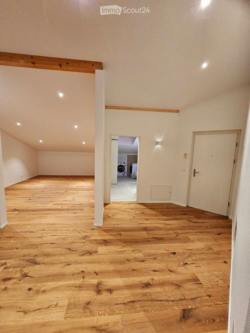 Empty room with wooden flooring, white walls, and recessed lighting. A doorway leads to a small laundry area with a washing machine and dryer.