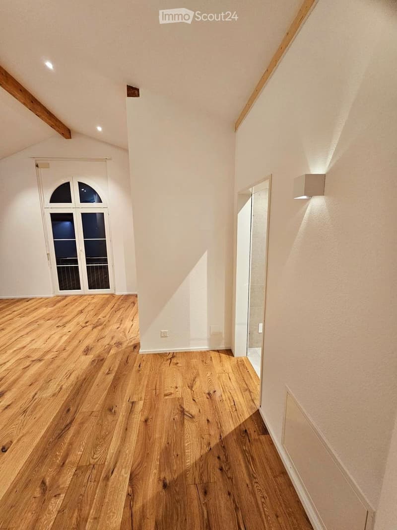Empty hallway with wooden flooring, white walls, and recessed lighting. Features a large arched window and a partially visible door frame on the right side.