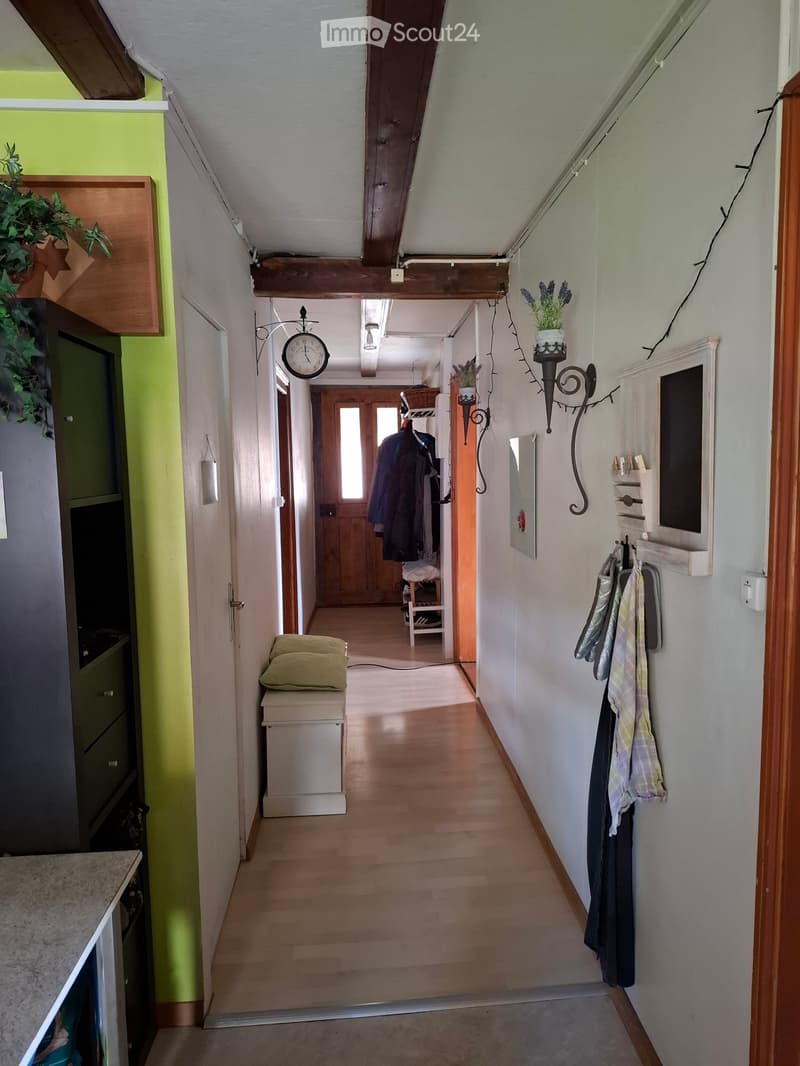 A narrow hallway with wooden flooring, white walls, and a slanted ceiling with exposed wooden beams. Features include a small bench with a cushion, a coat rack with hanging coats, a wall-mounted clock, and decorative wall-mounted planters with flowers. A