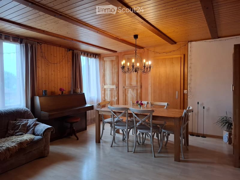 Cozy living and dining area with wooden walls and ceiling, featuring a sofa, piano, dining table with chairs, and a chandelier.
