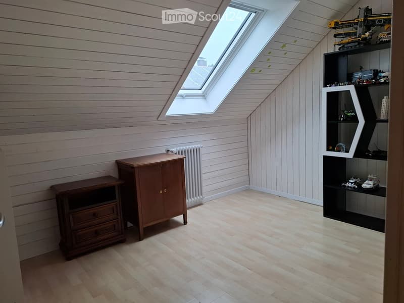 Bright attic room with wooden walls and floor, skylight windows, a small wooden cabinet, a shelf with toys, and a radiator.