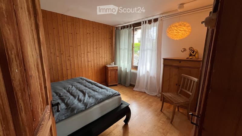 Cozy bedroom with wooden walls and floor, featuring a bed with a grey duvet, a wooden chair, a small nightstand, a wooden table with a decorative statue, and a hanging lamp.
