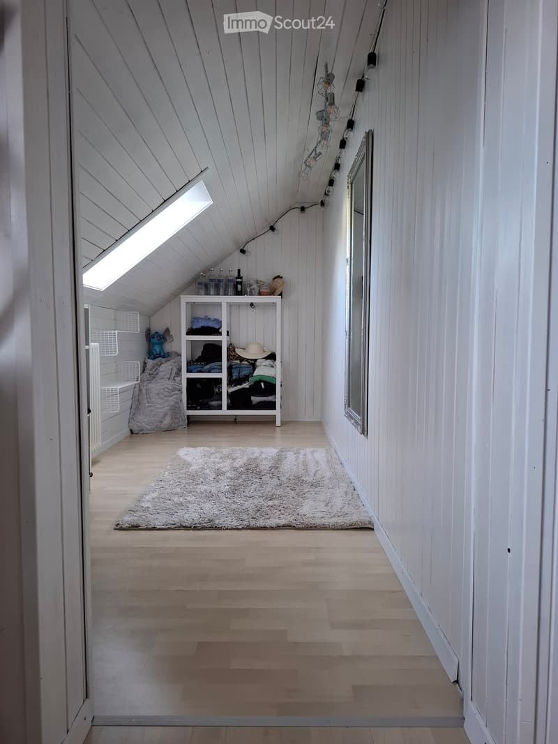 A small, cozy attic space with wooden walls and flooring, featuring a white shelf with folded clothes, a plush toy, and a hat, a large white rug, and a mirror on the wall.