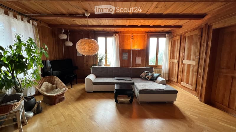 Cozy living room with wooden walls and ceiling, large windows, grey sectional sofa, coffee table, wicker chair, piano, and decorative lamps.