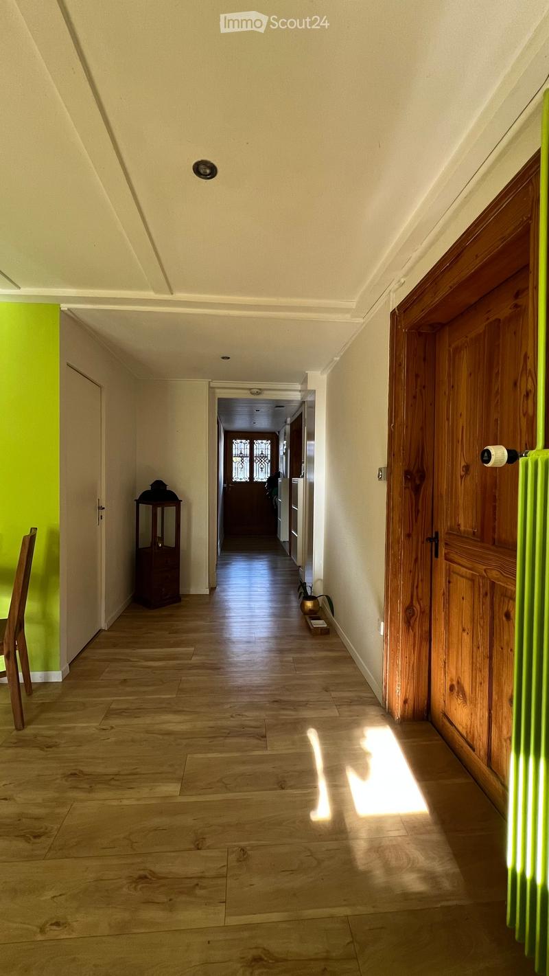 A long, narrow hallway with wooden flooring, white walls, and wooden doors on both sides. Natural light streams in from the end of the hallway, illuminating the space.