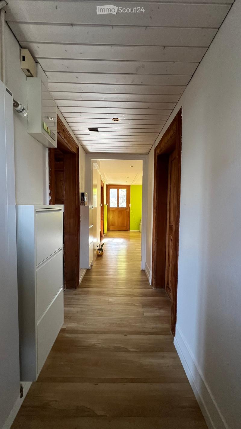 A long, narrow hallway with wooden flooring, white walls, and wooden doors on both sides. The ceiling is paneled with recessed lighting. A small decorative item is visible at the end of the hallway.