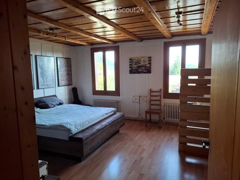 Bright bedroom with wooden floors, exposed wooden ceiling beams, large windows, bed with grey bedding, wooden chair, guitar, radiator, and artwork on walls.