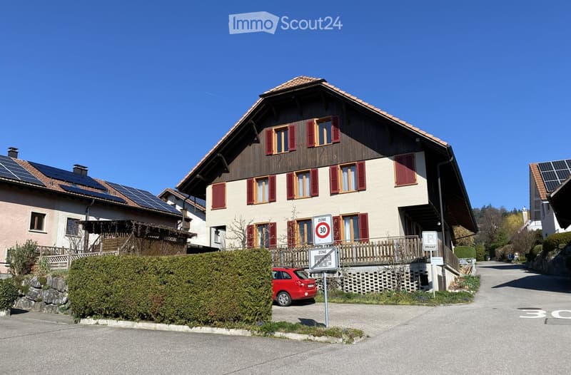 Two-story house with red shutters, solar panels on the roof, and a small balcony. A red car is parked in front, and there are speed limit signs visible.