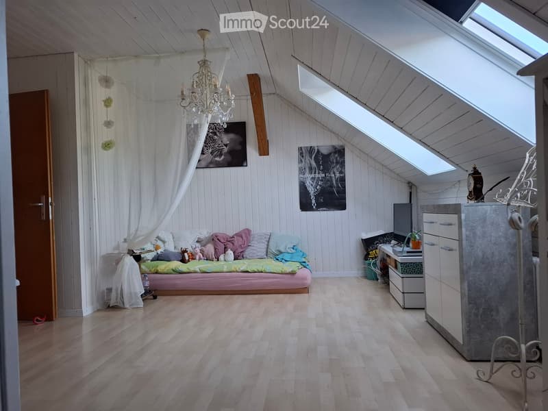 Bright attic bedroom with wooden floors, slanted ceilings, skylights, and a cozy sofa bed with colorful pillows and blankets. A chandelier hangs from the ceiling, and a small desk with a laptop is positioned near the window.